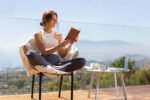 Mulher jovem sorridente, vestindo regata branca e calça legging cinza, sentada em poltrona confortável com as pernas cruzadas, lendo um livro em uma varanda de vidro com vista para montanhas, cidade e céu azul, transmitindo sensação de paz e bem-estar no contexto de despertar espiritual.