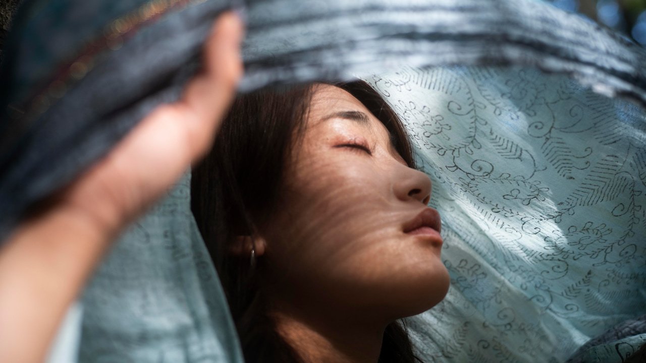 Mulher de olhos fechados com expressão serena, parcialmente coberta por um tecido azul claro translúcido com padrões florais, iluminada por luz natural filtrada, simbolizando tranquilidade e expansão da consciência após o despertar espiritual