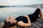 Mulher jovem deitada de costas sobre um deck de madeira, usando vestido preto e sandálias claras, olhando serenamente para o céu, com um lago calmo e montanhas ao fundo, transmitindo tranquilidade e introspecção associadas ao despertar espiritual