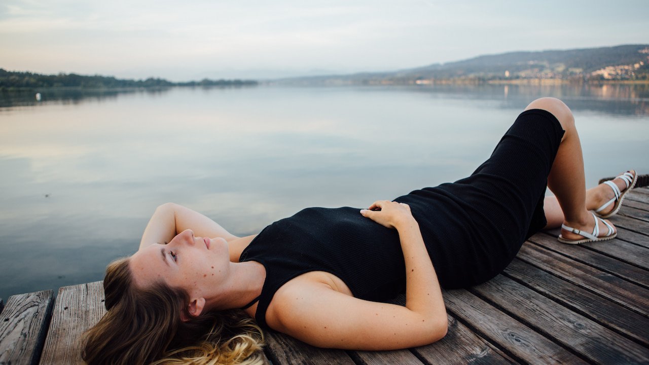 Mulher jovem deitada de costas sobre um deck de madeira, usando vestido preto e sandálias claras, olhando serenamente para o céu, com um lago calmo e montanhas ao fundo, transmitindo tranquilidade e introspecção associadas ao despertar espiritual