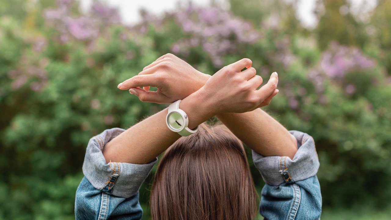 Pessoa de costas com cabelos castanhos, vestindo jaqueta jeans e erguendo os braços cruzados sobre a cabeça, usando relógio de pulso branco com mostrador verde claro, em ambiente externo com fundo desfocado de vegetação e flores lilás, simbolizando reflexão sobre o tempo do despertar espiritual